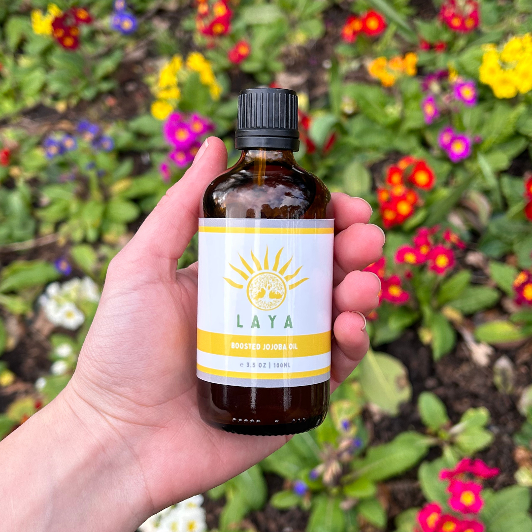 Hand holding Laya's Boosted Jojoba oil, over a bed of red, purple, white & yellow flowers.