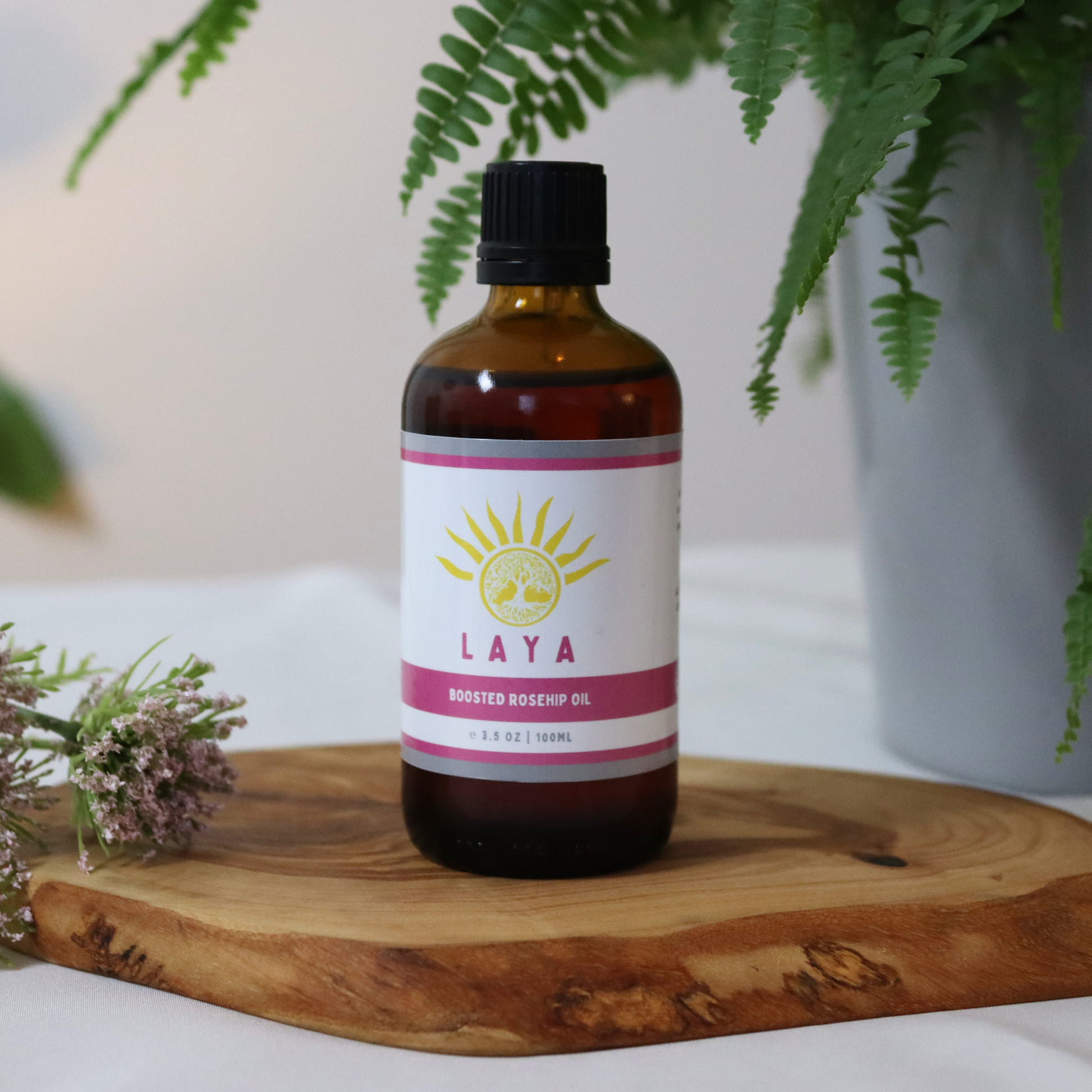 Boosted rosehip oil by Laya in 100ml in amber glass bottle. Displayed on wood next to plant and purple flower.