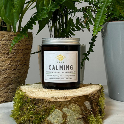 Natural calming candle by Laya in amber glass jar, displayed on wood & set against peaceful backdrop.